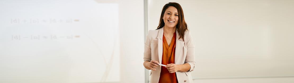 A teacher stands and smiles in front of a whiteboard